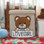 Beige cotton rope storage basket with embroidered teddy bear and Lovegirl A beautifully crafted beige cotton rope storage basket sits in a cozy nursery setting, showcasing its embroidered teddy bear motif and intricate Lovegirl Pattern. The removable lid is slightly open, revealing soft baby blankets inside, while the sturdy handles and natural texture of the eco-friendly cotton rope highlight its durability. The basket’s compact 10x10 inch size makes it ideal for shelves, corners, or as a decorative accent. Designed for safety and style, it blends seamlessly with neutral or pastel nursery themes. Pattern, removable lid, and handles – 10x10 inches (25x25 cm) nursery hamper